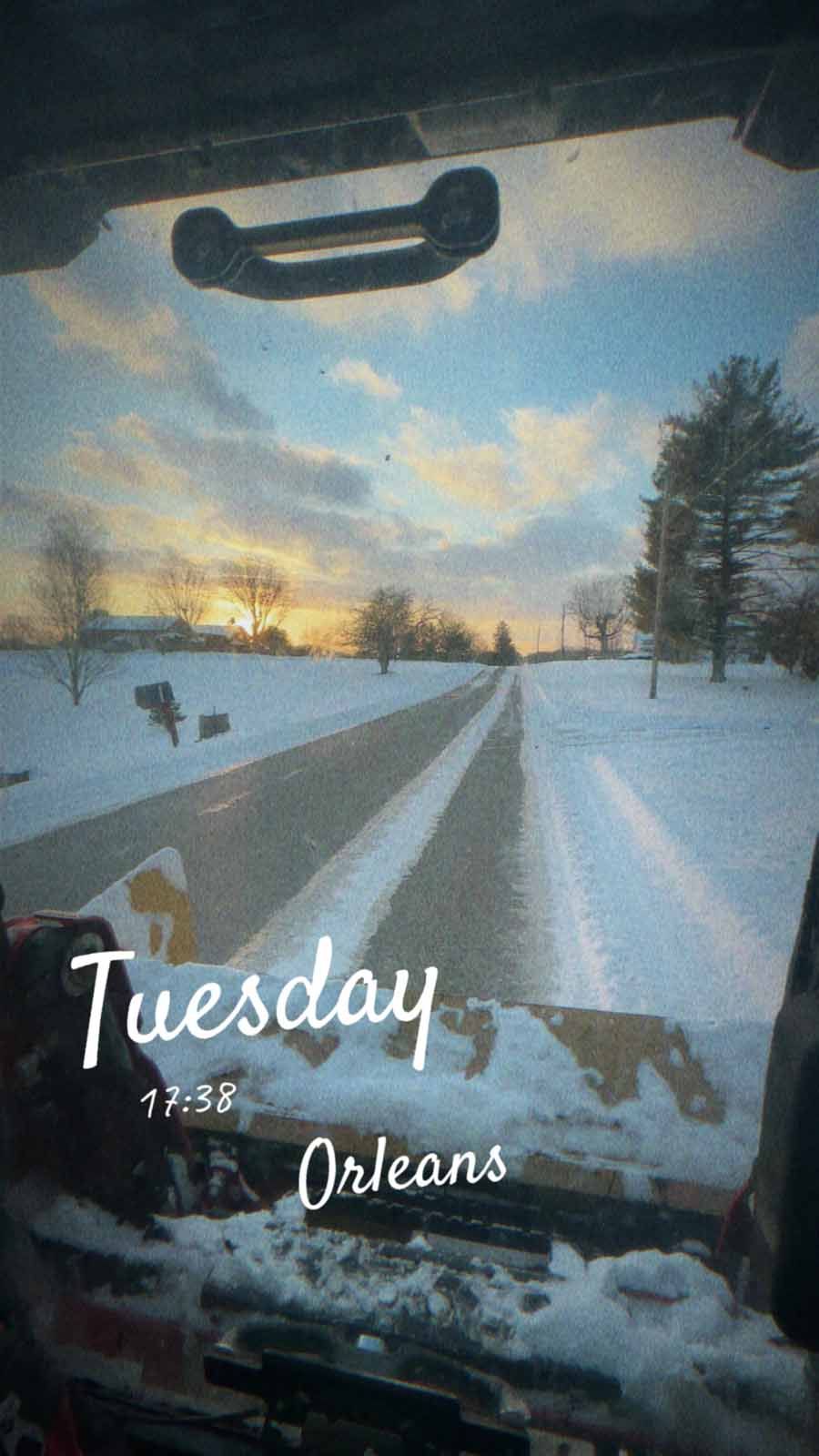 A snowy road viewed through a vehicle windshield at sunset, with 
