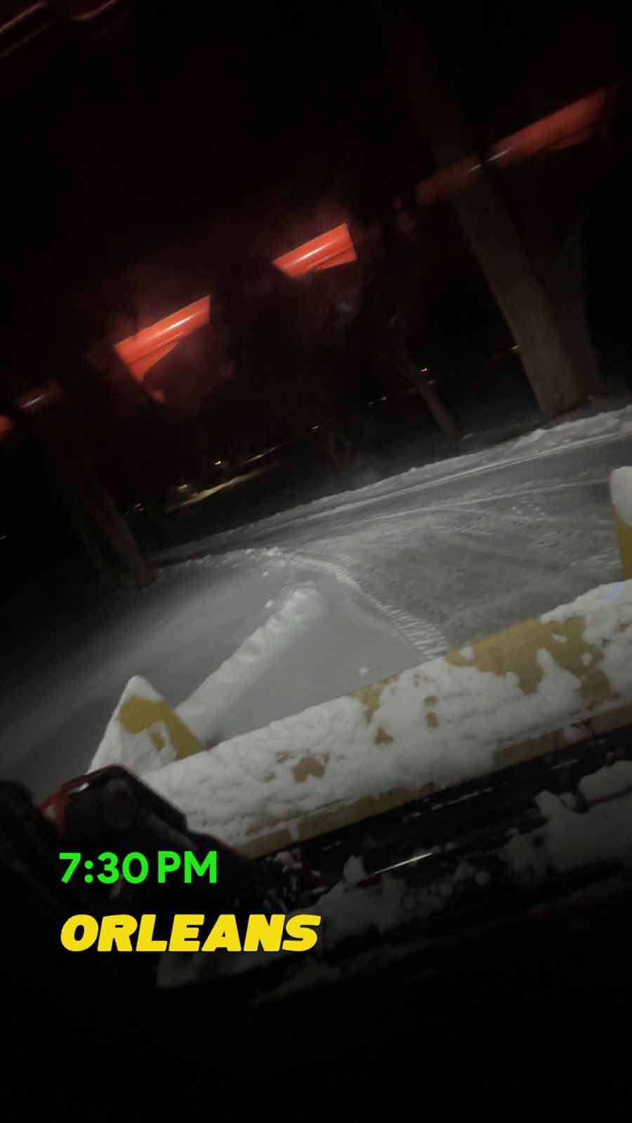 A snowplow blade clears a dark, snow-covered road at night, with 