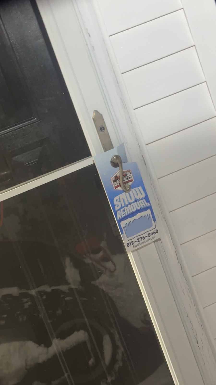 A light blue snow removal advertisement door hanger is hanging from the handle of a white storm door.
