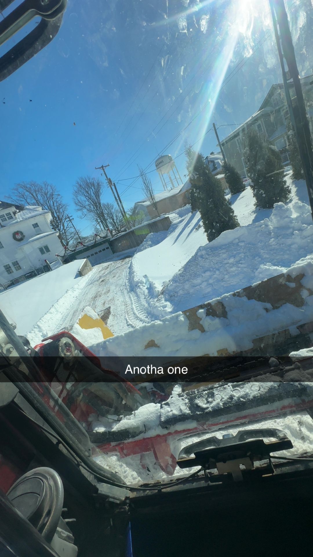 View from inside a plow truck cab on a bright, sunny day, showing cleared snow and a nearby building.