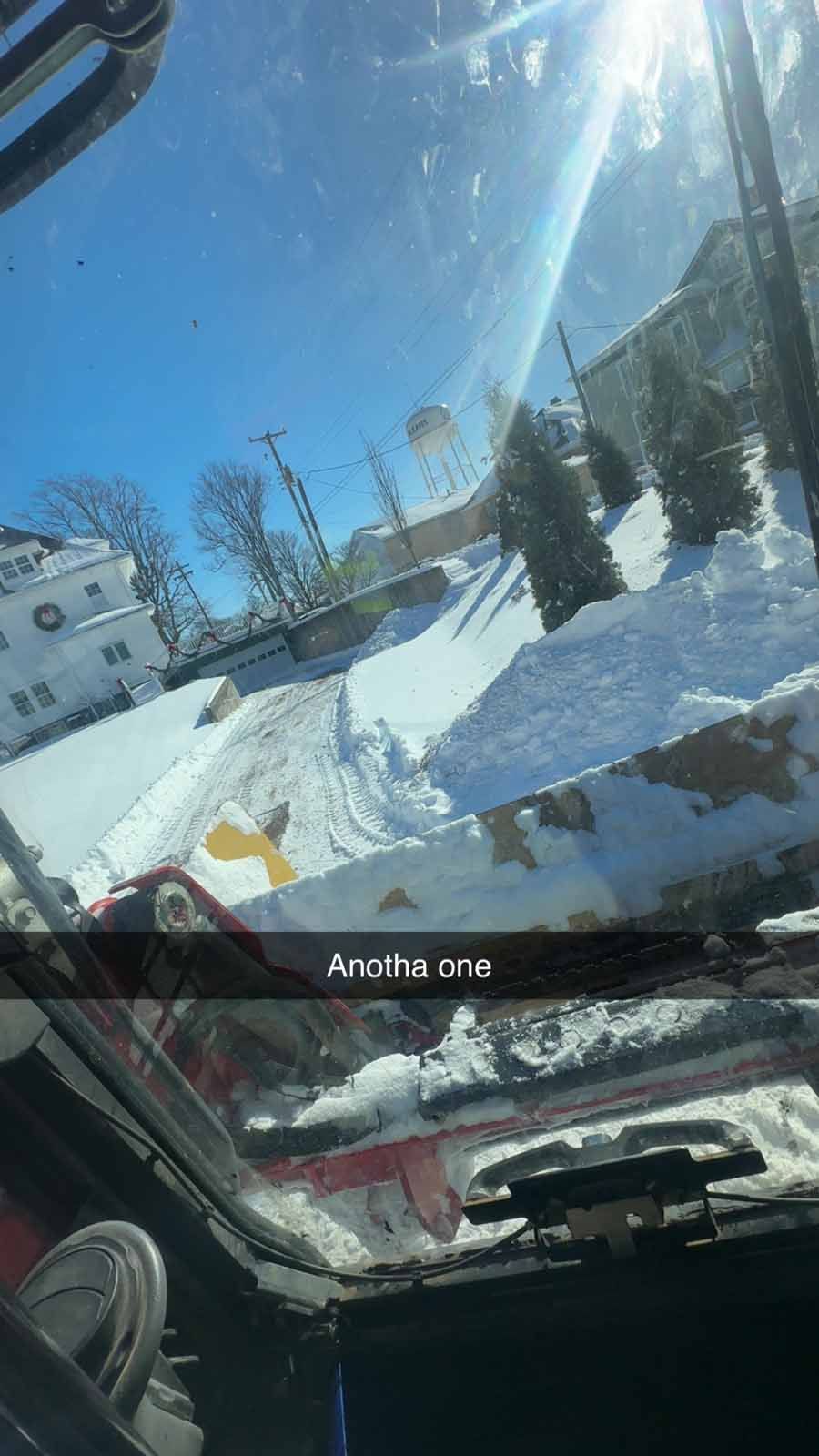 A view from inside a snowplow cab, showing a snow-covered street and buildings on a bright, sunny day.