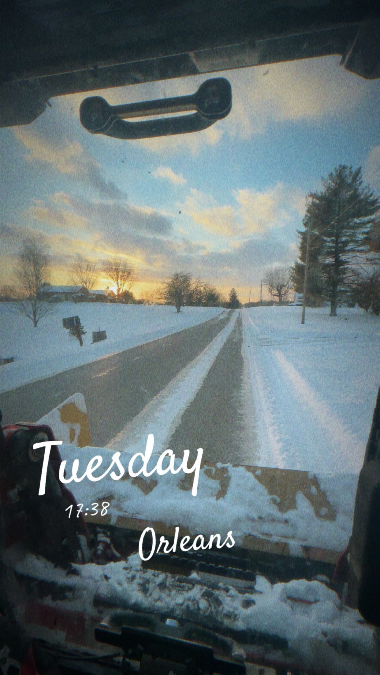 A view from inside a snowplow cab looking out onto a snow-covered road at sunset in Orleans, with a Tuesday timestamp.