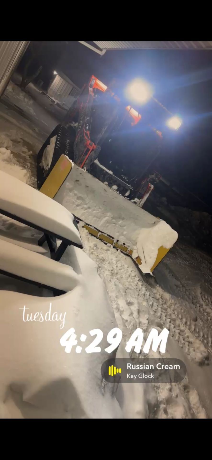 A tractor with a snow plow at night, seen next to a snow-covered picnic table, with an overlay reading 4:29 AM on Tuesday.