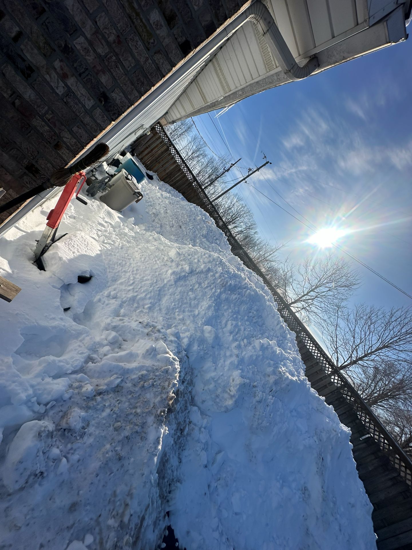 A snowy backyard with a tall snowdrift piled against a house siding and a wooden fence under a bright, sunny sky.