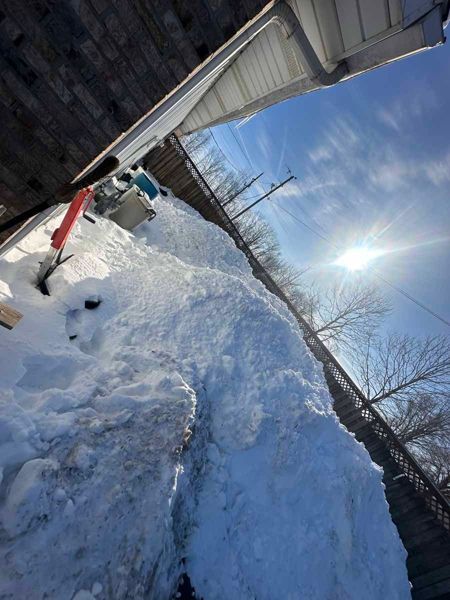 A large pile of snow accumulates against a residential fence beneath a bright, sunny sky.