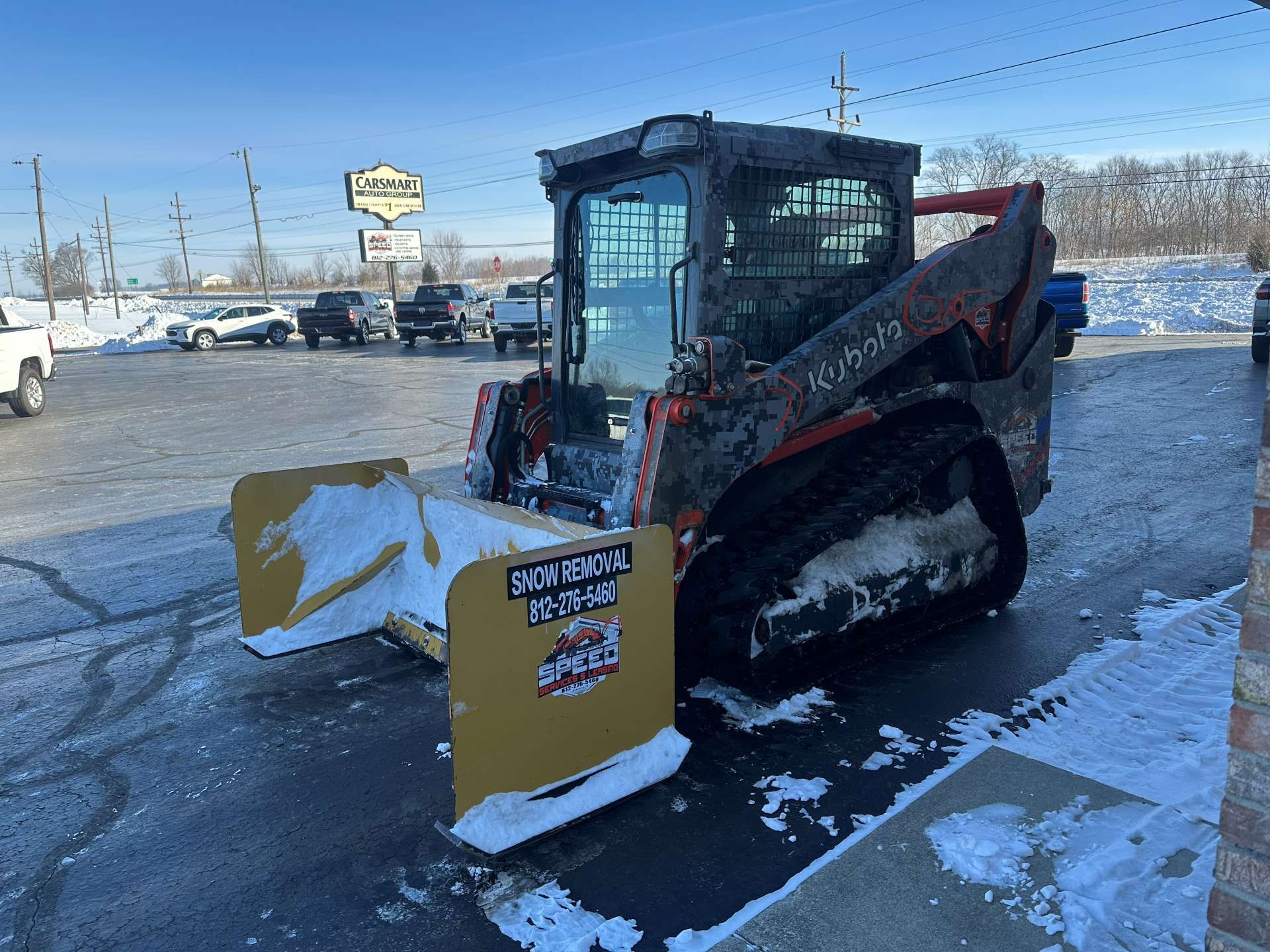 A Kubota tracked skid steer with a large yellow snow pusher attachment parked on a snowy, paved lot.