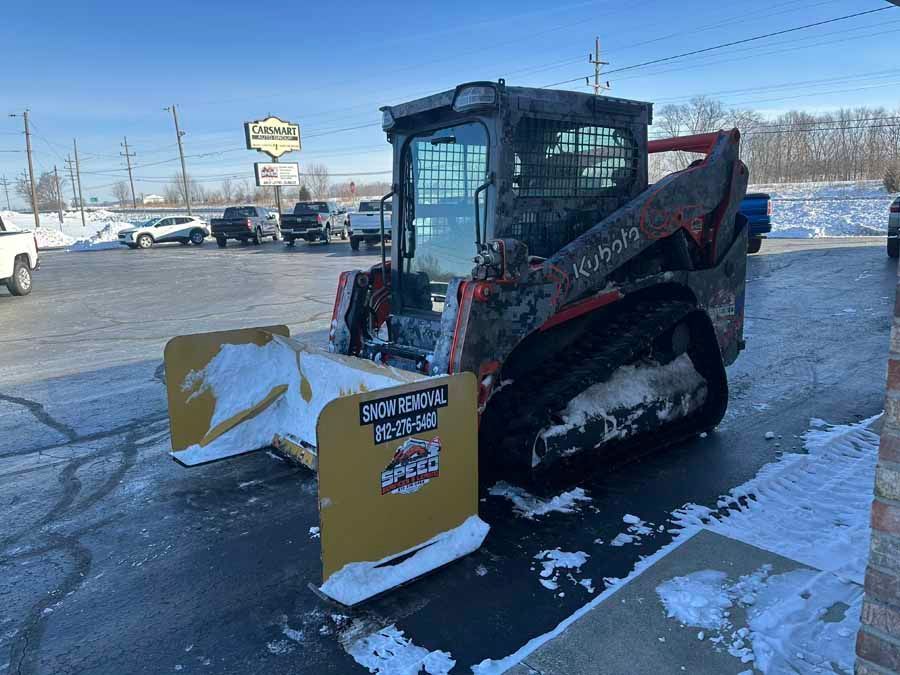 An orange Kubota track loader with a large yellow snow pusher attachment parked in a snow-covered parking lot.