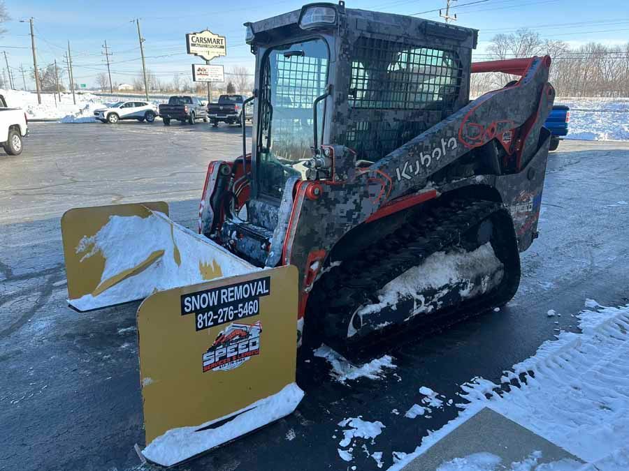 A Kubota compact track loader with a large yellow snow pusher attachment parked in a snowy lot.
