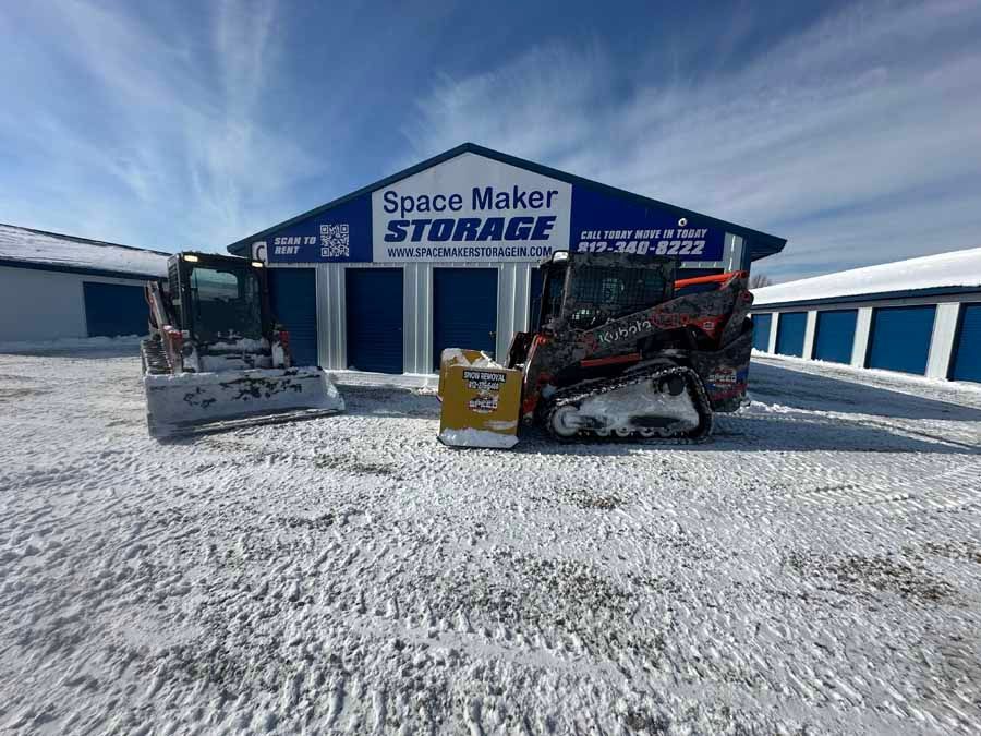 Two skid steers with snowplows parked in front of a blue and white Space Maker Storage building on a snowy lot.