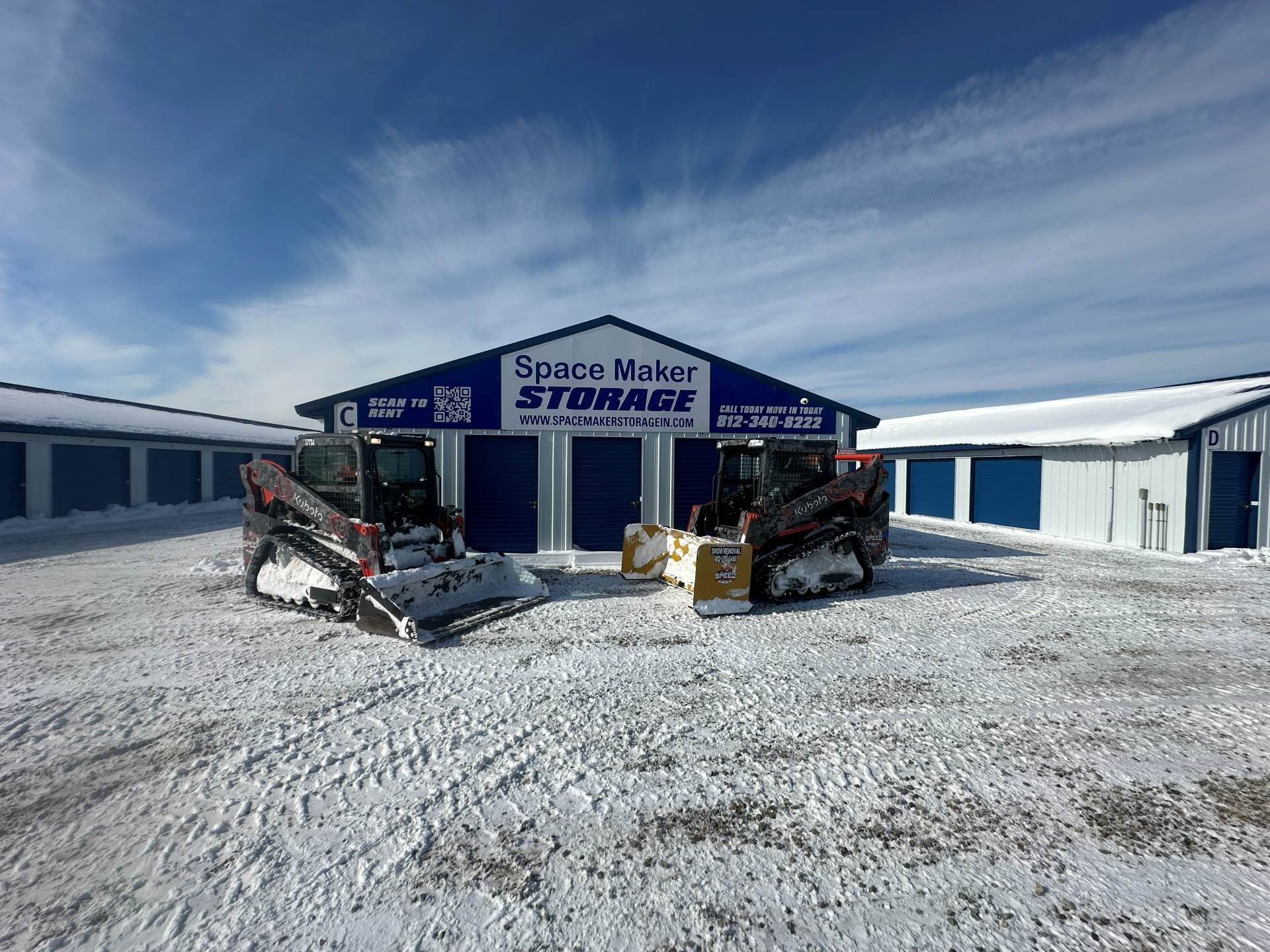 Two skid steers parked in front of a blue and white Space Maker Storage building on a snow-covered lot under a sunny sky.