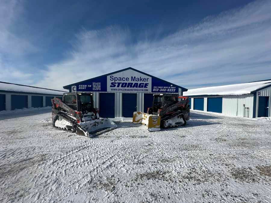 Two skid-steer loaders parked in the snow in front of a blue and white Space Maker Storage facility.