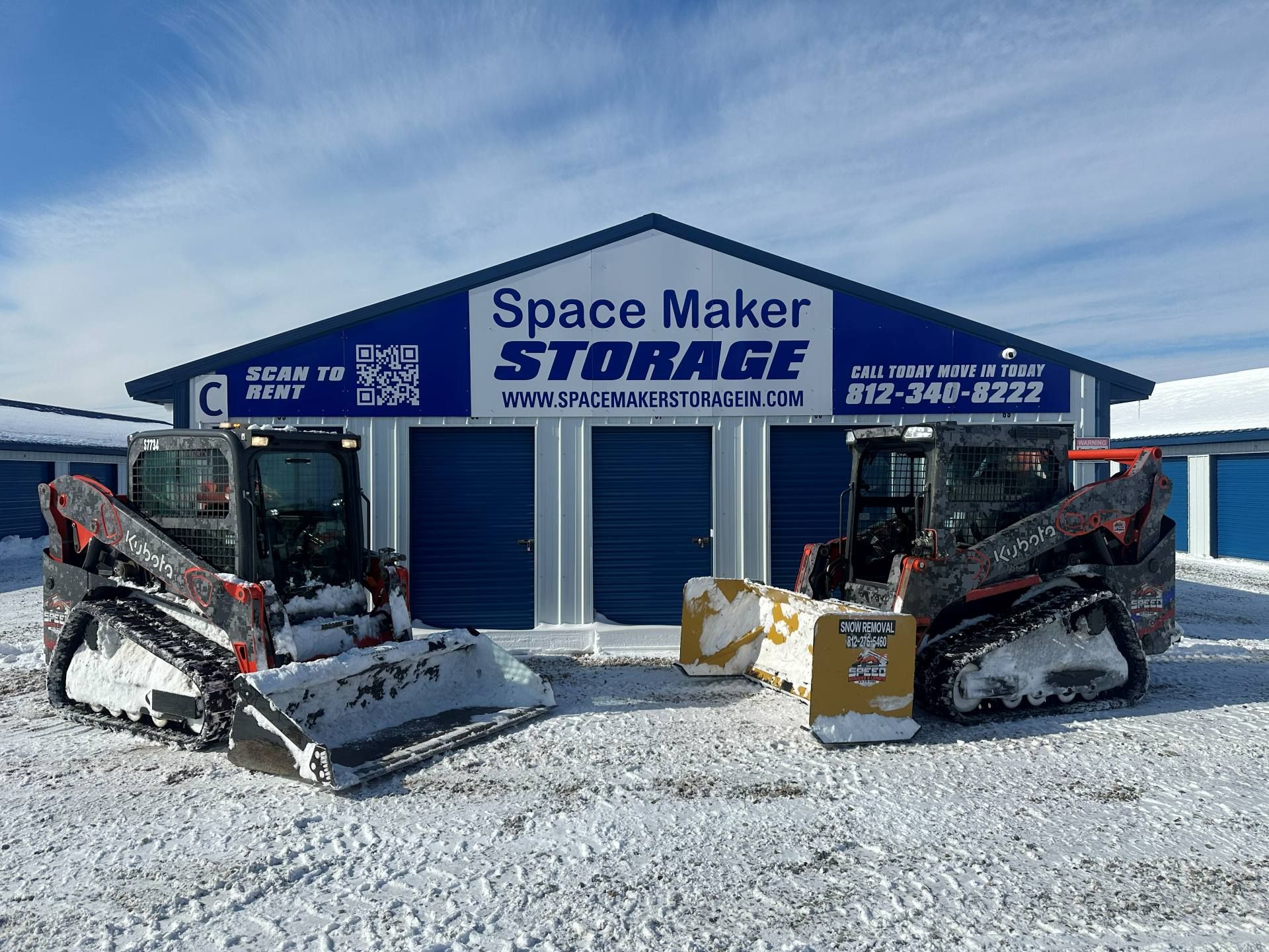 Two skid steer loaders with snow blades parked in front of a blue and white Space Maker Storage building in the snow.