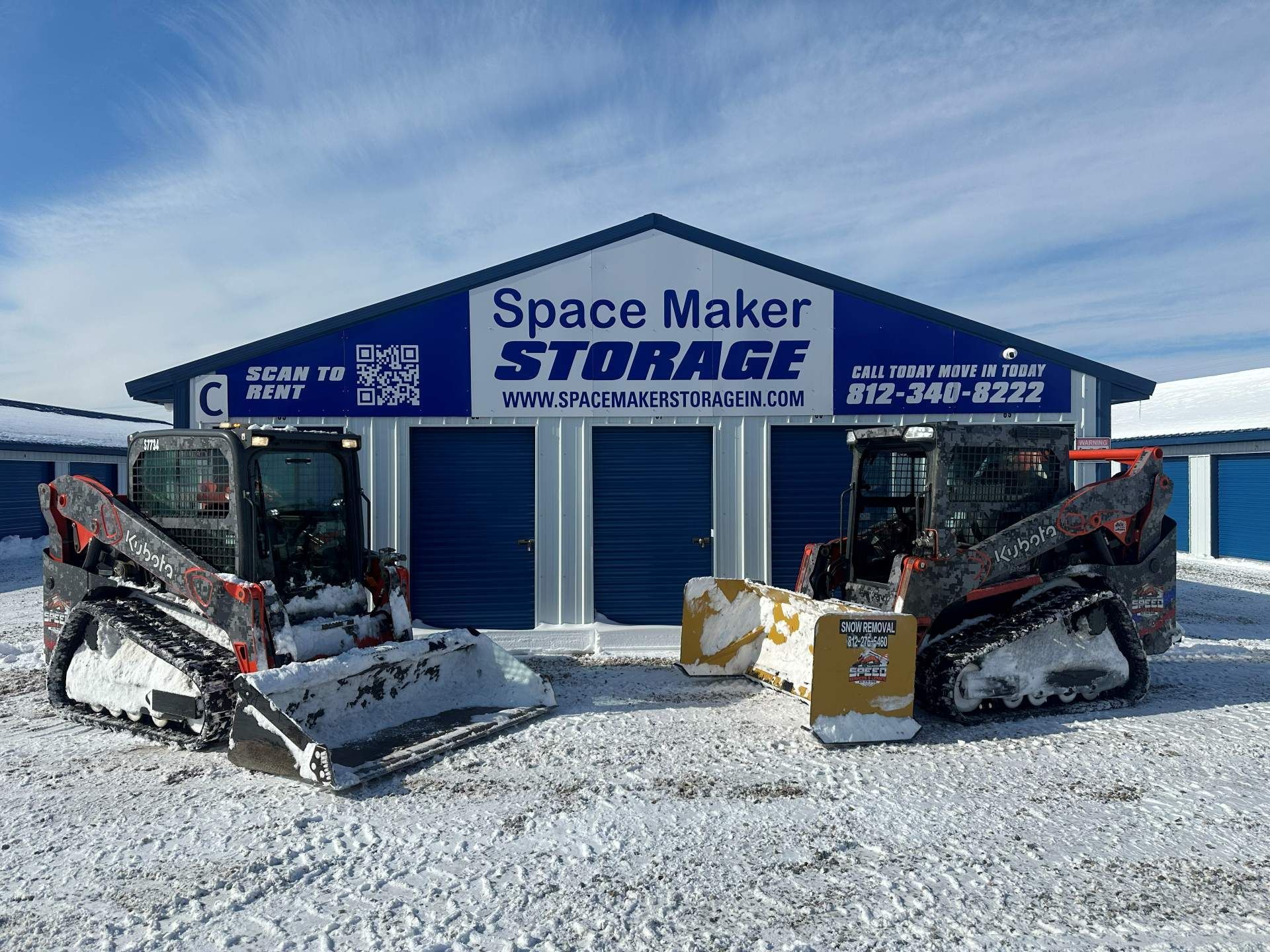 Two skid steer loaders with snow blades parked in front of a blue and white Space Maker Storage building in the snow.