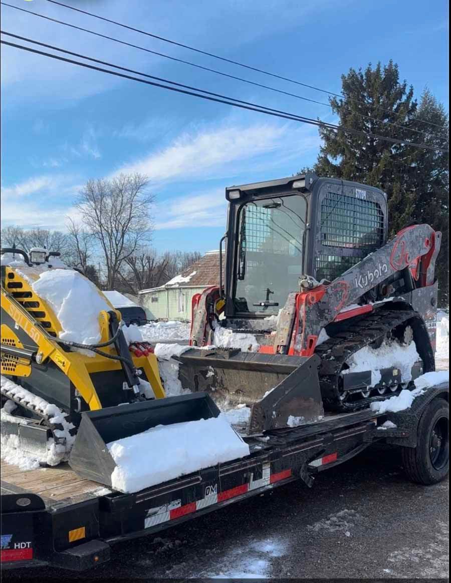 A yellow and a red compact track loader are parked on a flatbed trailer outdoors, covered in patches of snow.