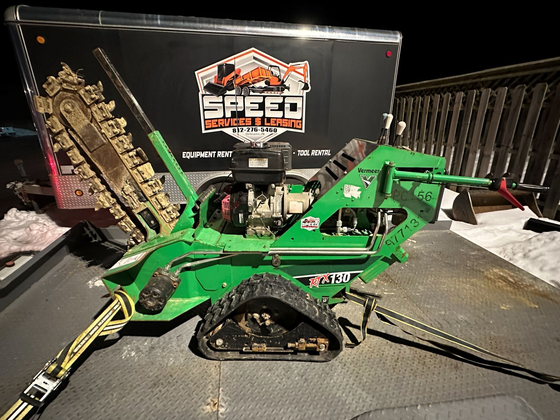 A bright green walk-behind trencher with black tracks secured by tie-down straps on a trailer at night.