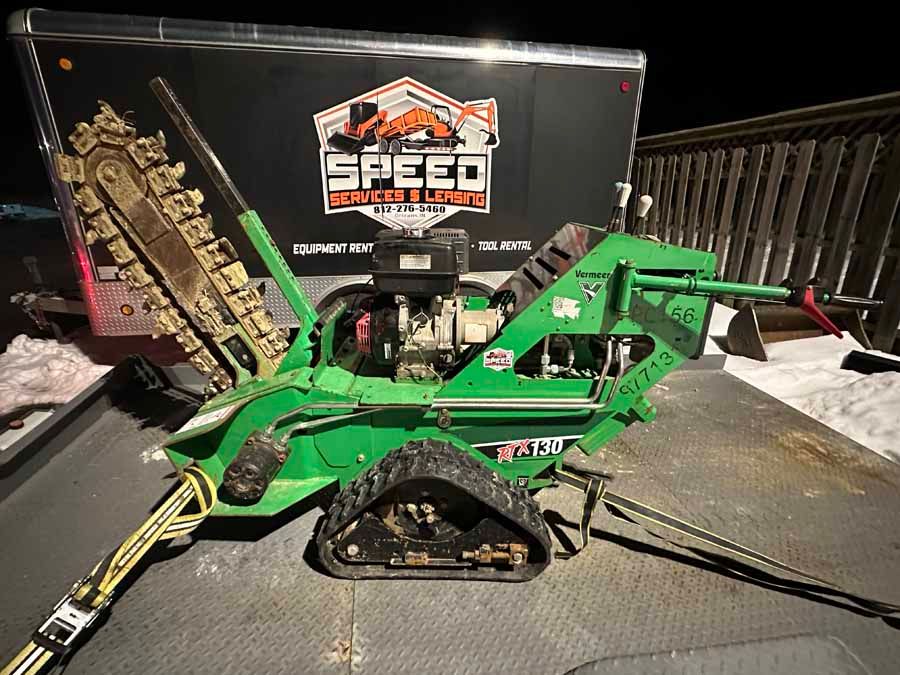 A green tracked trencher machine secured on a trailer at night, with a branded black trailer in the background.