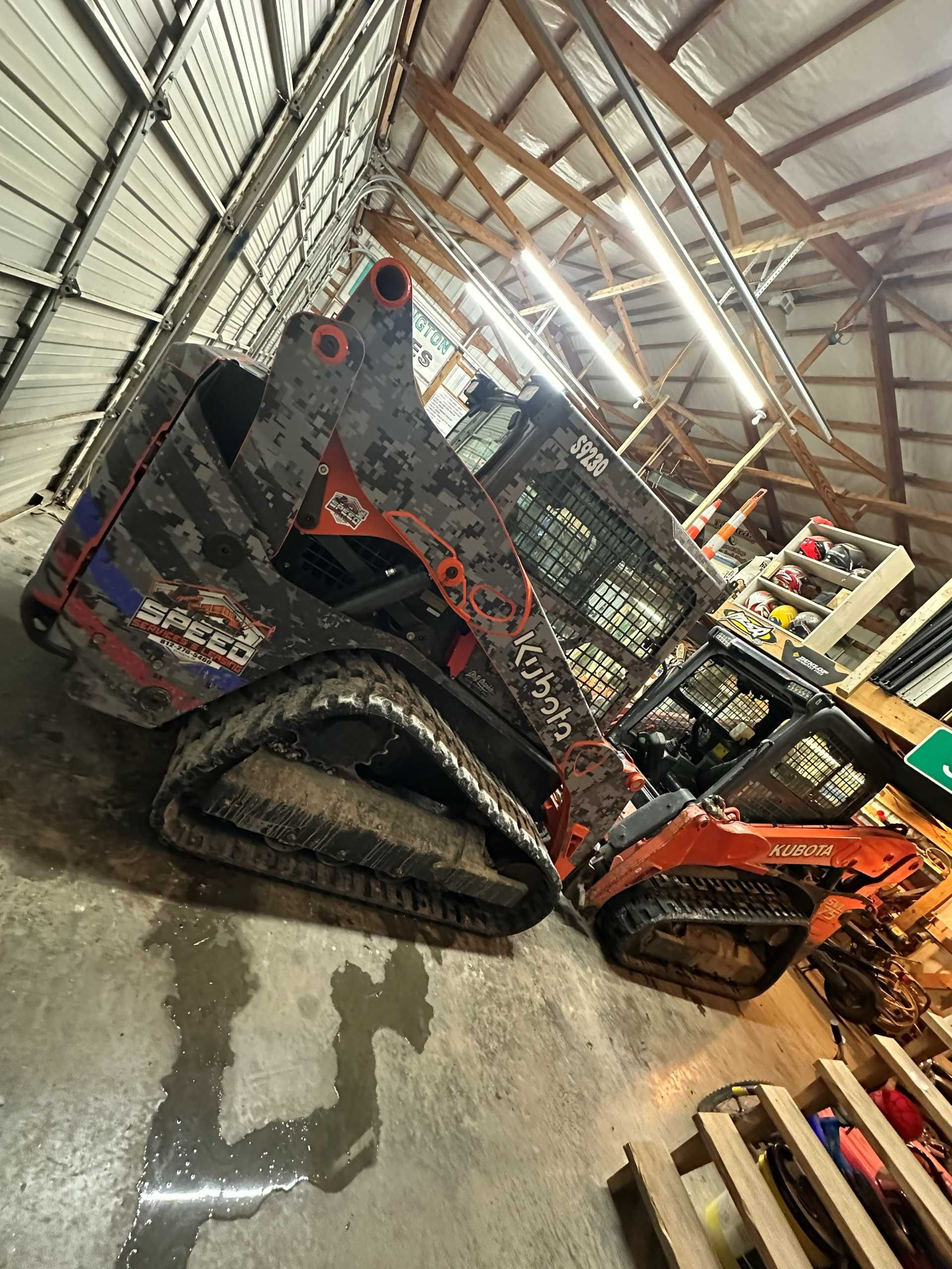 An orange Kubota track loader sits parked on a concrete floor inside a workshop with wooden rafters and overhead lights.