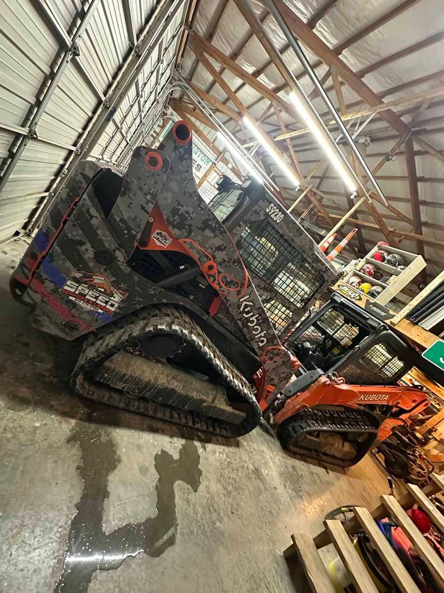 An orange Kubota compact track loader with a custom American flag wrap parked inside a workshop with wooden rafters.