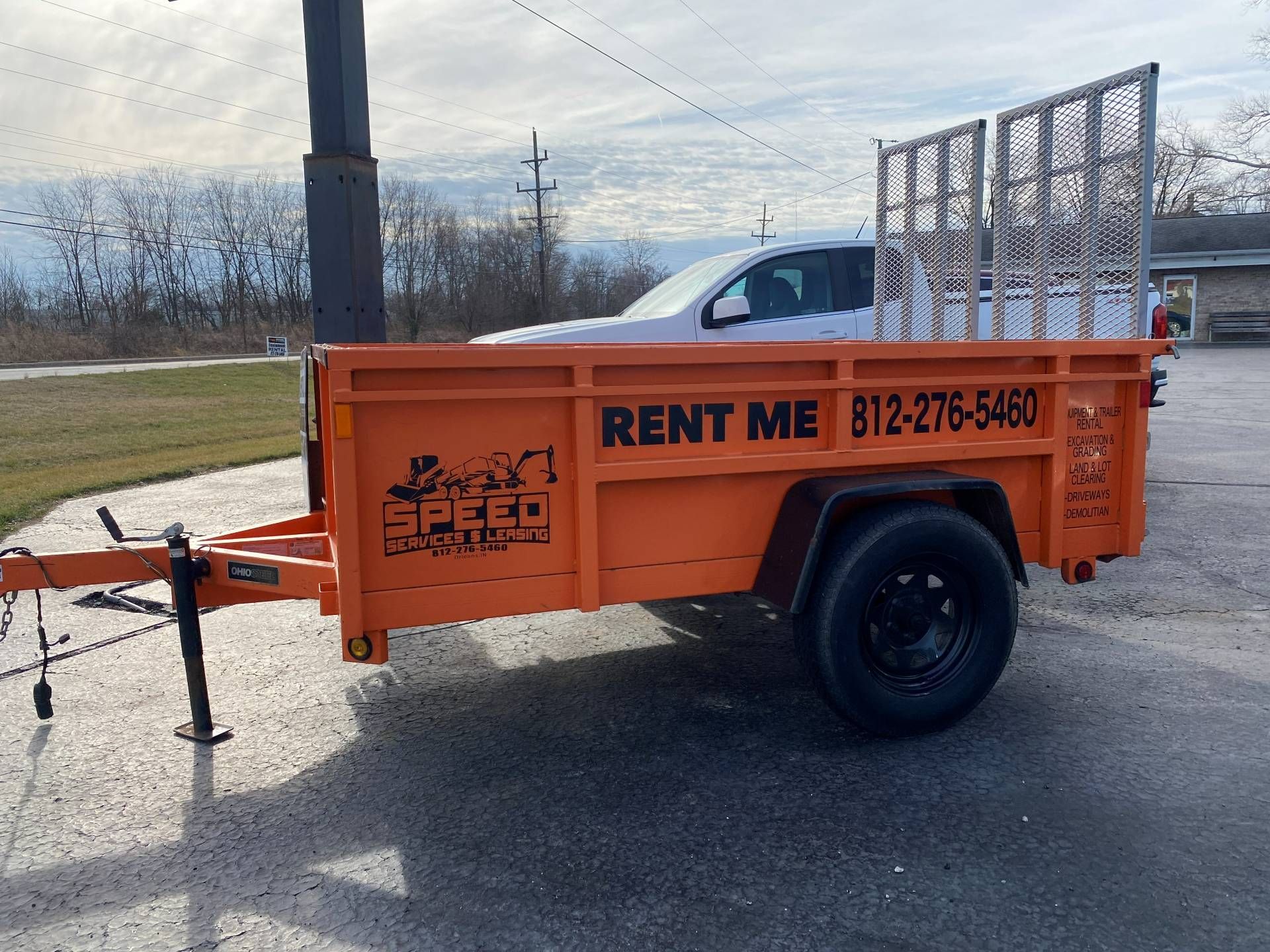 An orange rental utility trailer with a mesh gate sits on a gravel lot with a white pickup truck in the background.