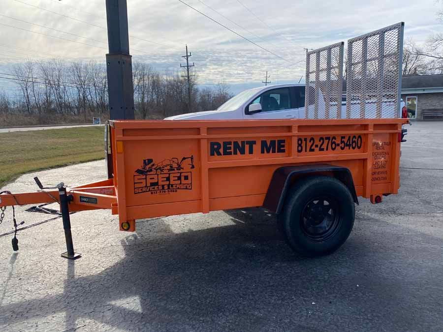 A bright orange rental utility trailer with a mesh gate sits in a gravel lot on a sunny day.
