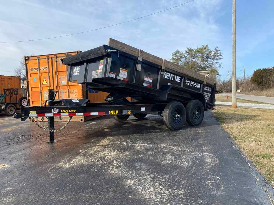 A black dump trailer hitched to a vehicle, parked on asphalt next to an orange shipping container under a blue sky.