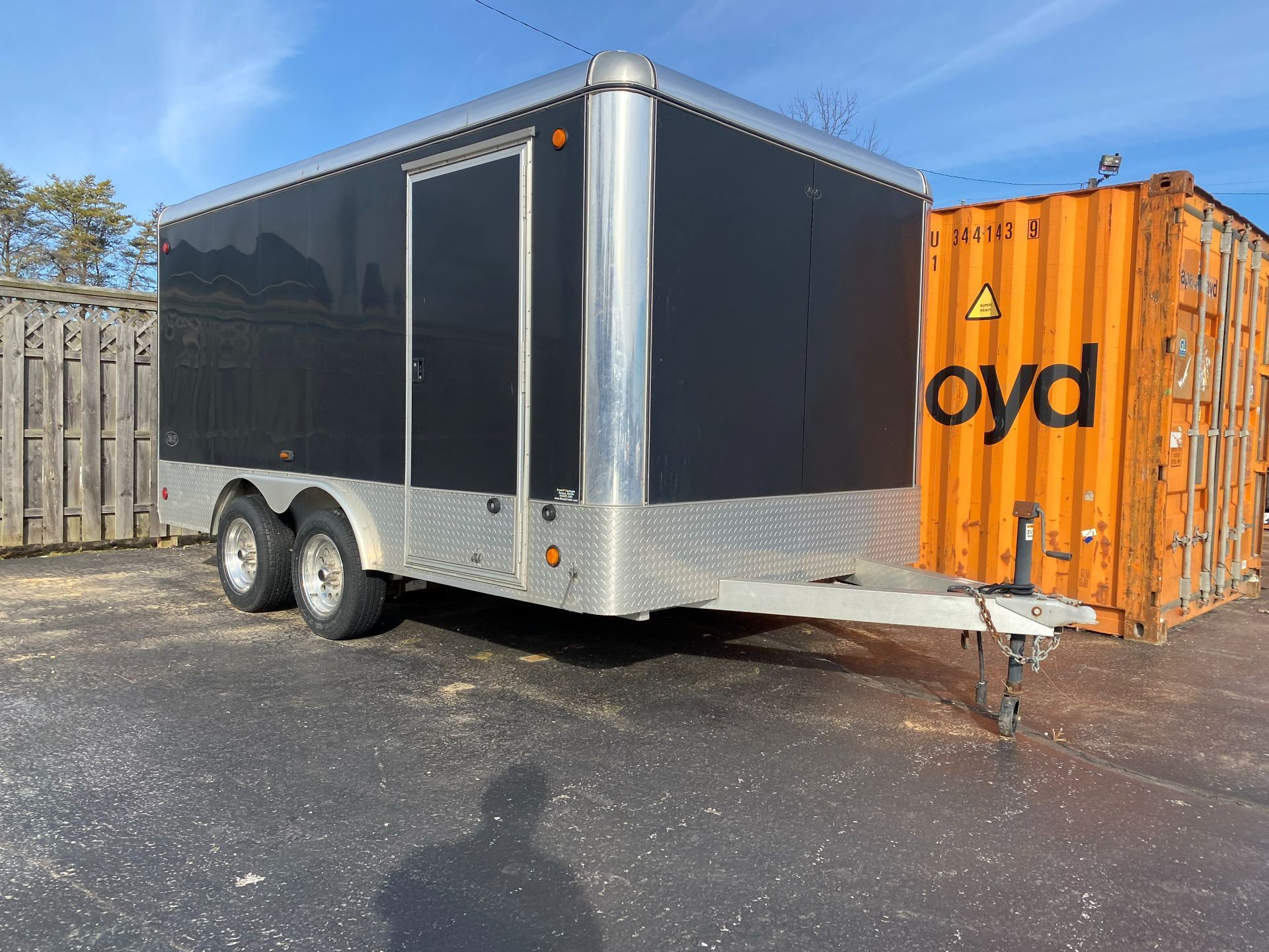A black tandem-axle enclosed trailer parked outdoors next to an orange shipping container on a sunny day.
