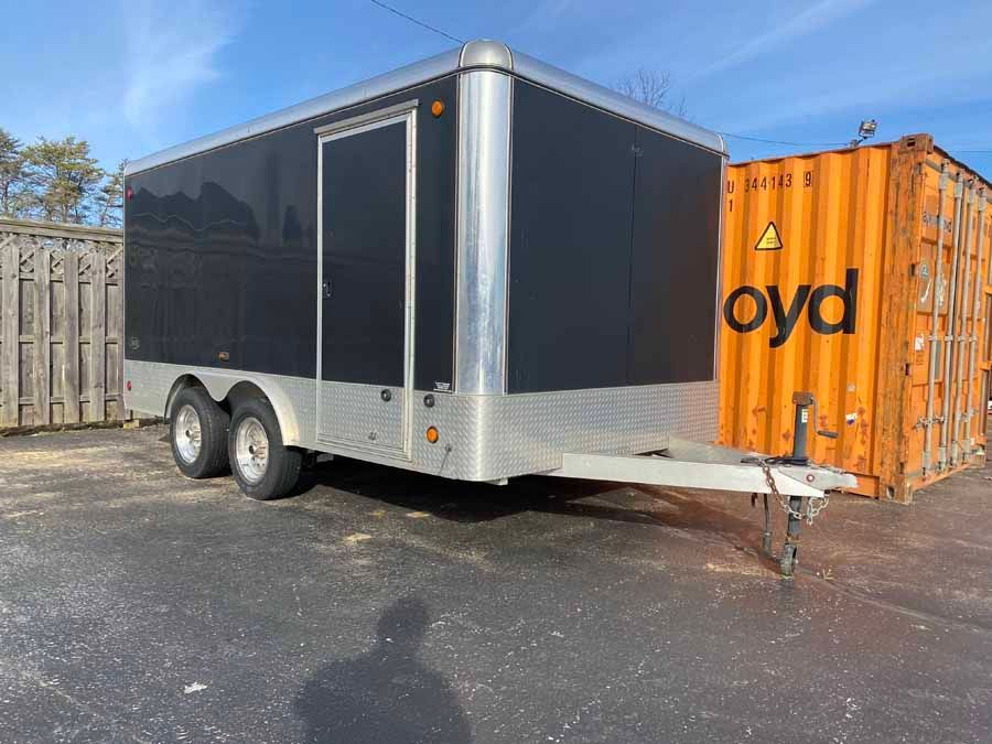 A black and silver enclosed cargo trailer parked outdoors next to an orange shipping container on a sunny day.