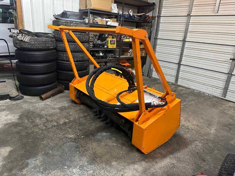 A bright orange hydraulic flail mower attachment for heavy equipment, sitting on a concrete floor in a garage.