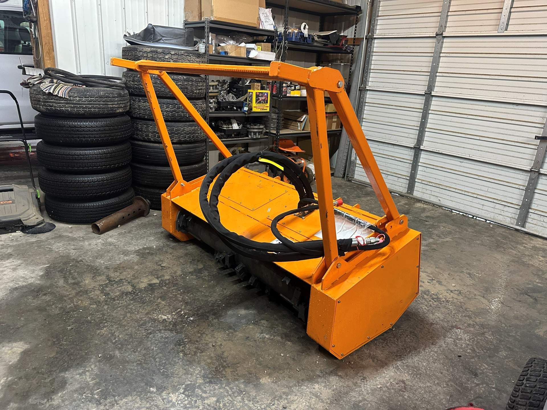 A bright orange industrial flail mower attachment for a tractor or skid steer sits on a concrete garage floor.