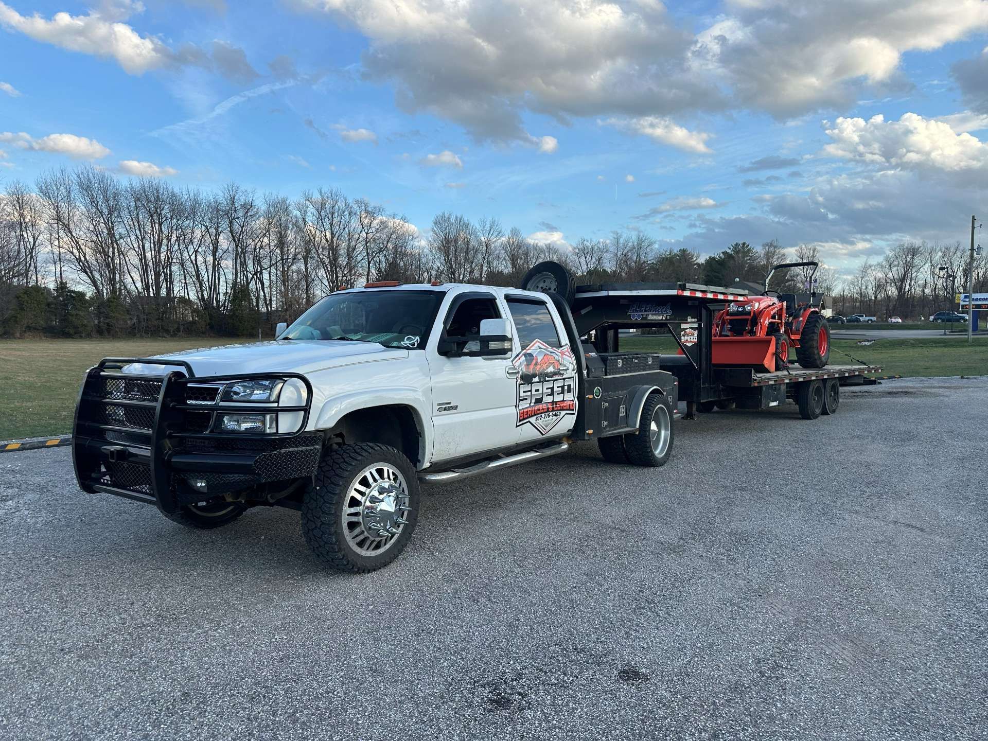 A white pickup truck with a black brush guard and large tires towing a flatbed trailer with an orange tractor.