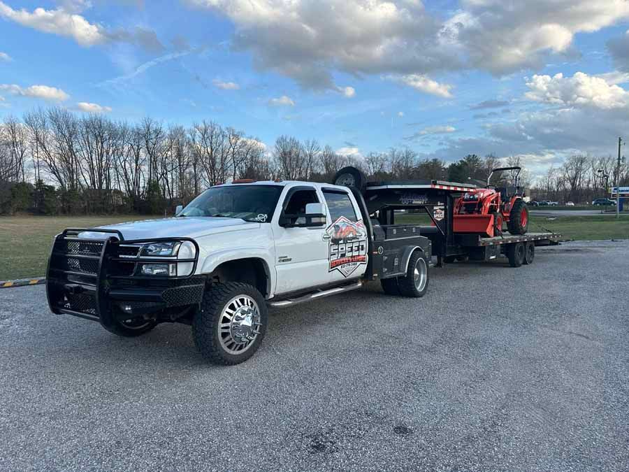 A white Chevrolet dually pickup truck with a large black grille guard pulling a trailer loaded with an orange tractor.