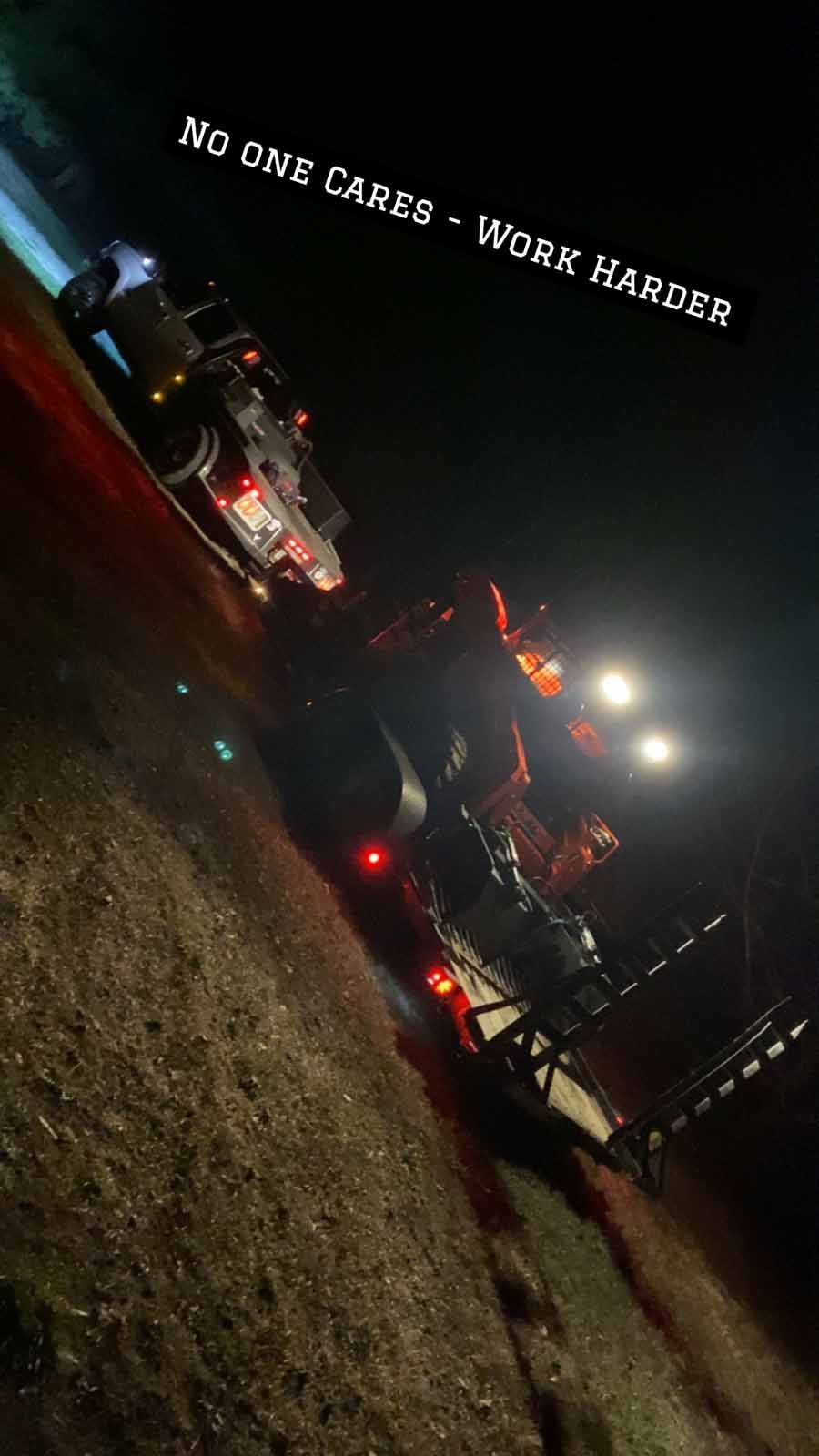 A truck towing a trailer with machinery at night under bright work lights, with the text 