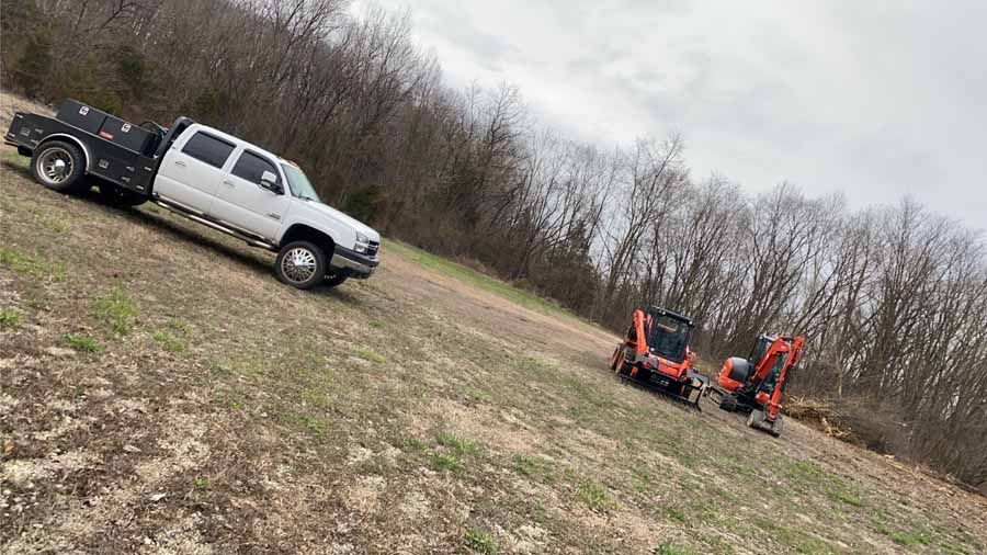 A white pickup truck with a flatbed and two orange construction vehicles parked in a grassy field near a forest.