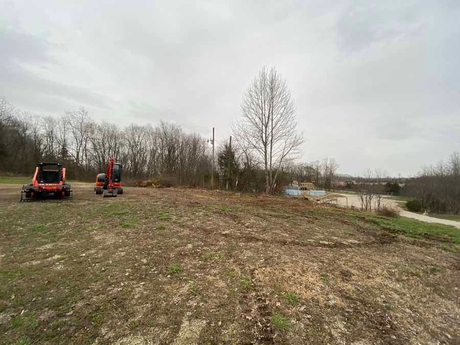 Two orange construction vehicles sit on a grassy, cleared field with trees and a pale, cloudy sky in the background.