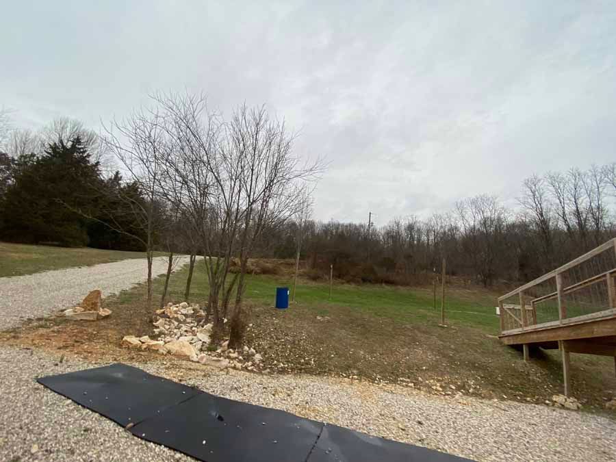 A gravel driveway leads toward a grassy area with a lone blue barrel, nearby trees, and a wooden ramp on the right.