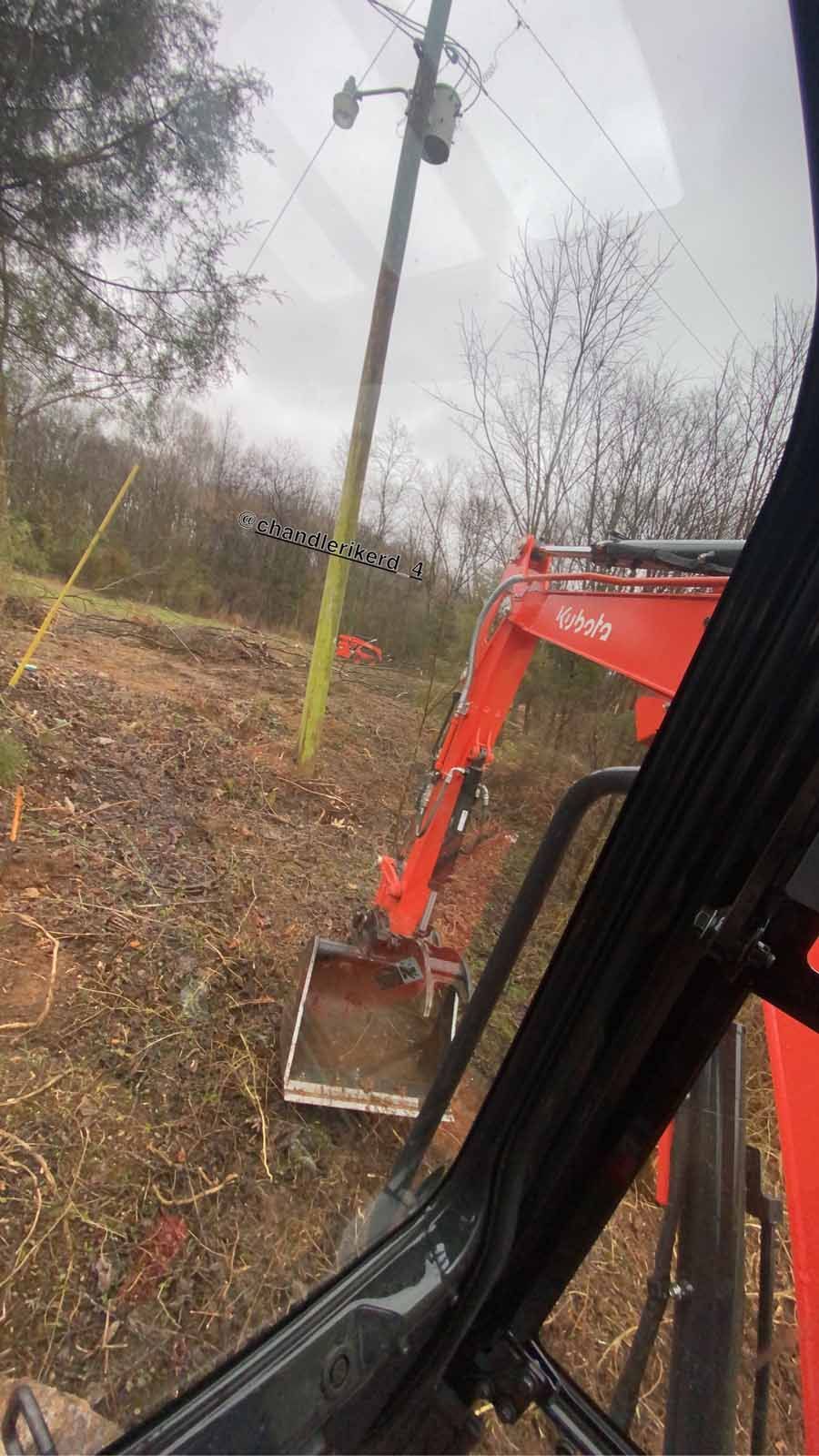 An orange Kubota excavator arm and bucket sit on bare, dirt ground near a wooden utility pole.