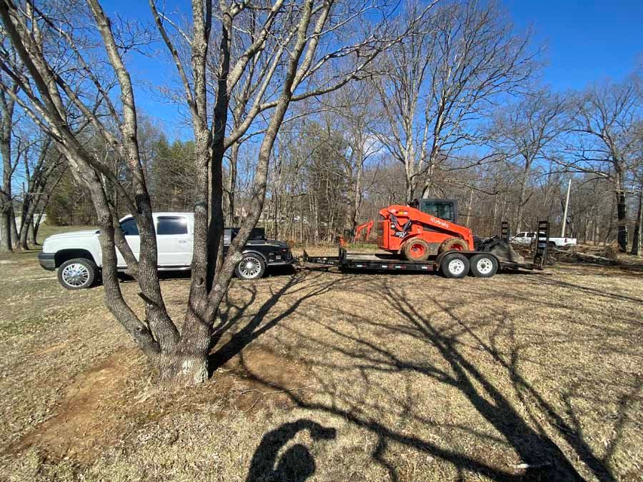 A white pickup truck towing a flatbed trailer with an orange skid-steer loader on it, parked in a sunny, wooded field.