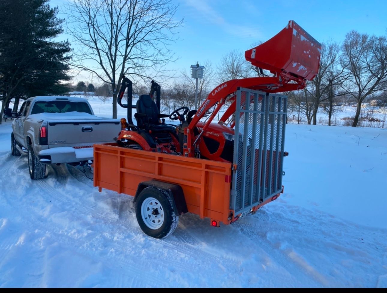 A pickup truck pulling an orange trailer carrying a Kubota tractor on a snowy, open landscape at dusk.