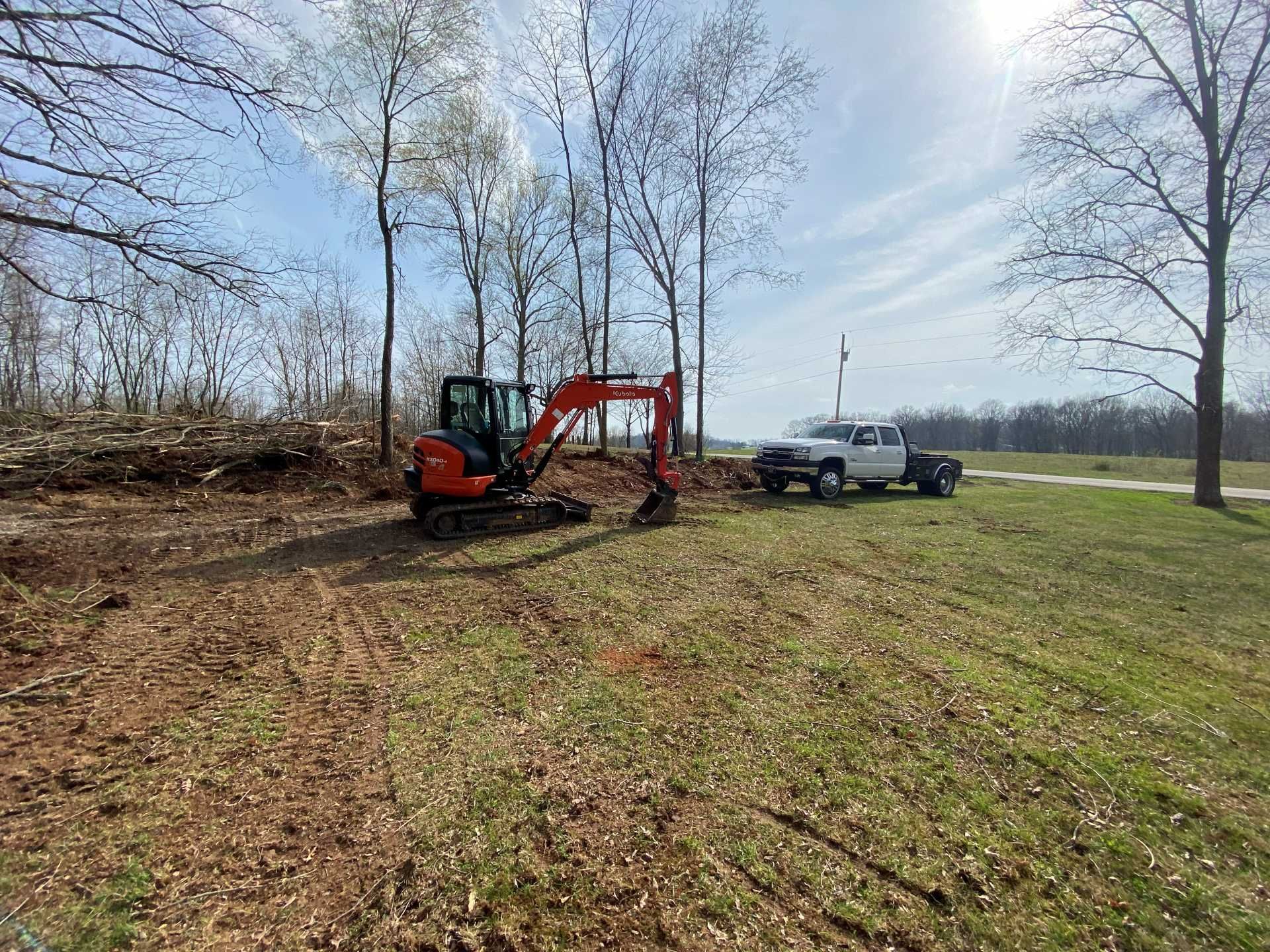 An orange mini excavator sits on a dirt lot next to a white pickup truck, with bare trees in the background under a blue sky.