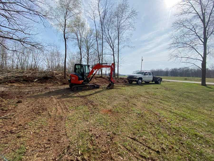 A red compact excavator sits in a clearing next to a white pickup truck on a grassy lot with trees in the background.
