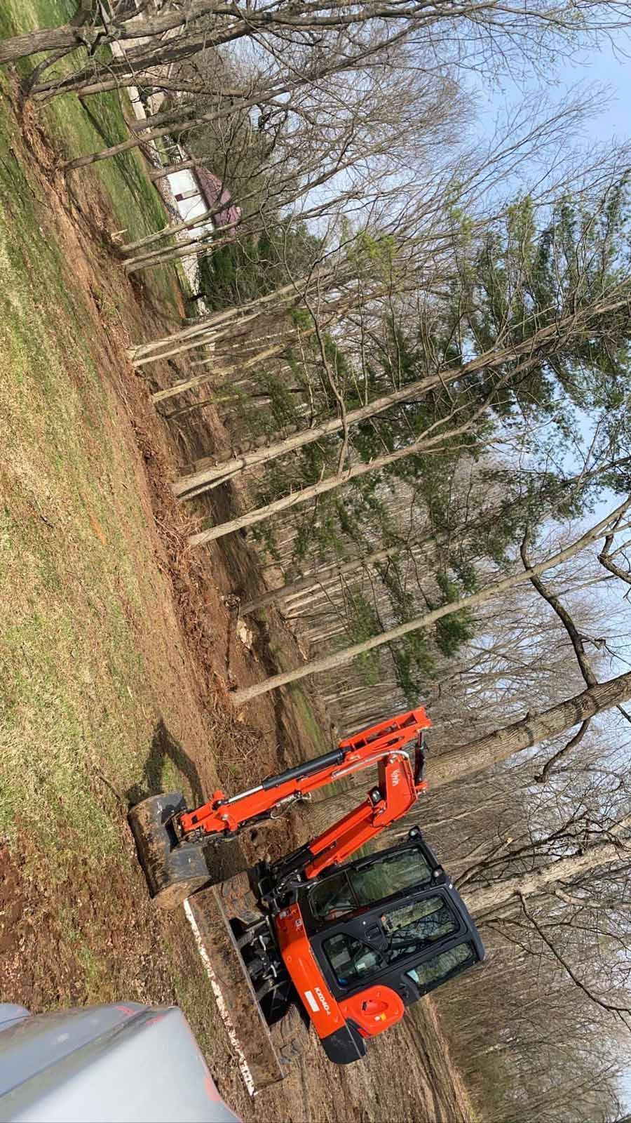 An orange excavator sits on a dirt lot cleared of brush next to a treeline in a residential area.