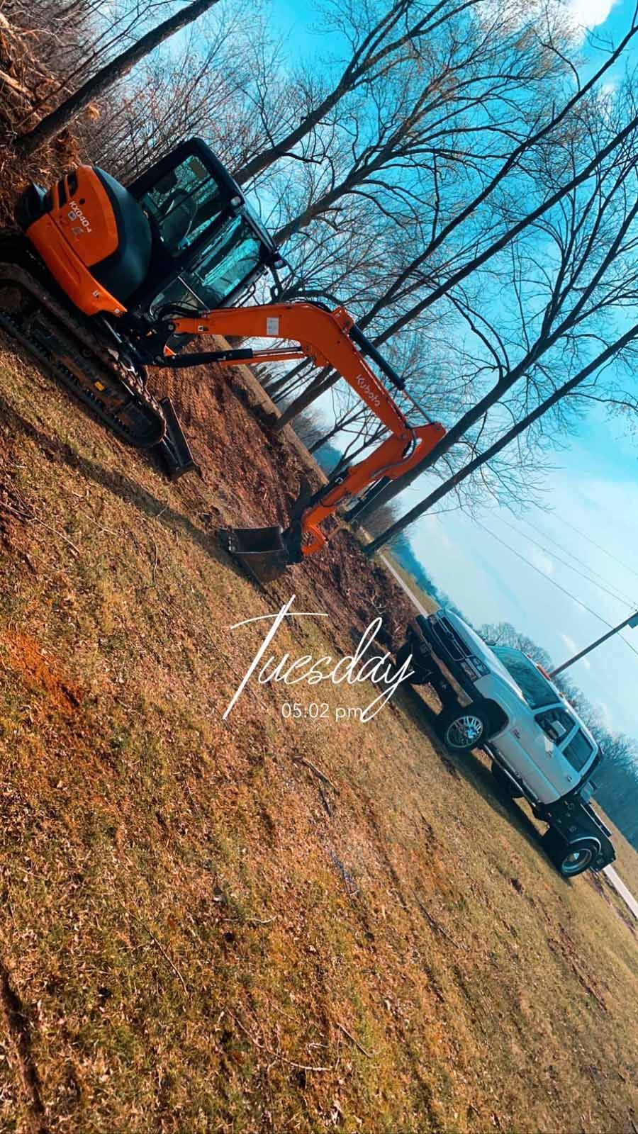 An orange excavator parked next to a white pickup truck on a grassy, wooded hillside on a bright day.