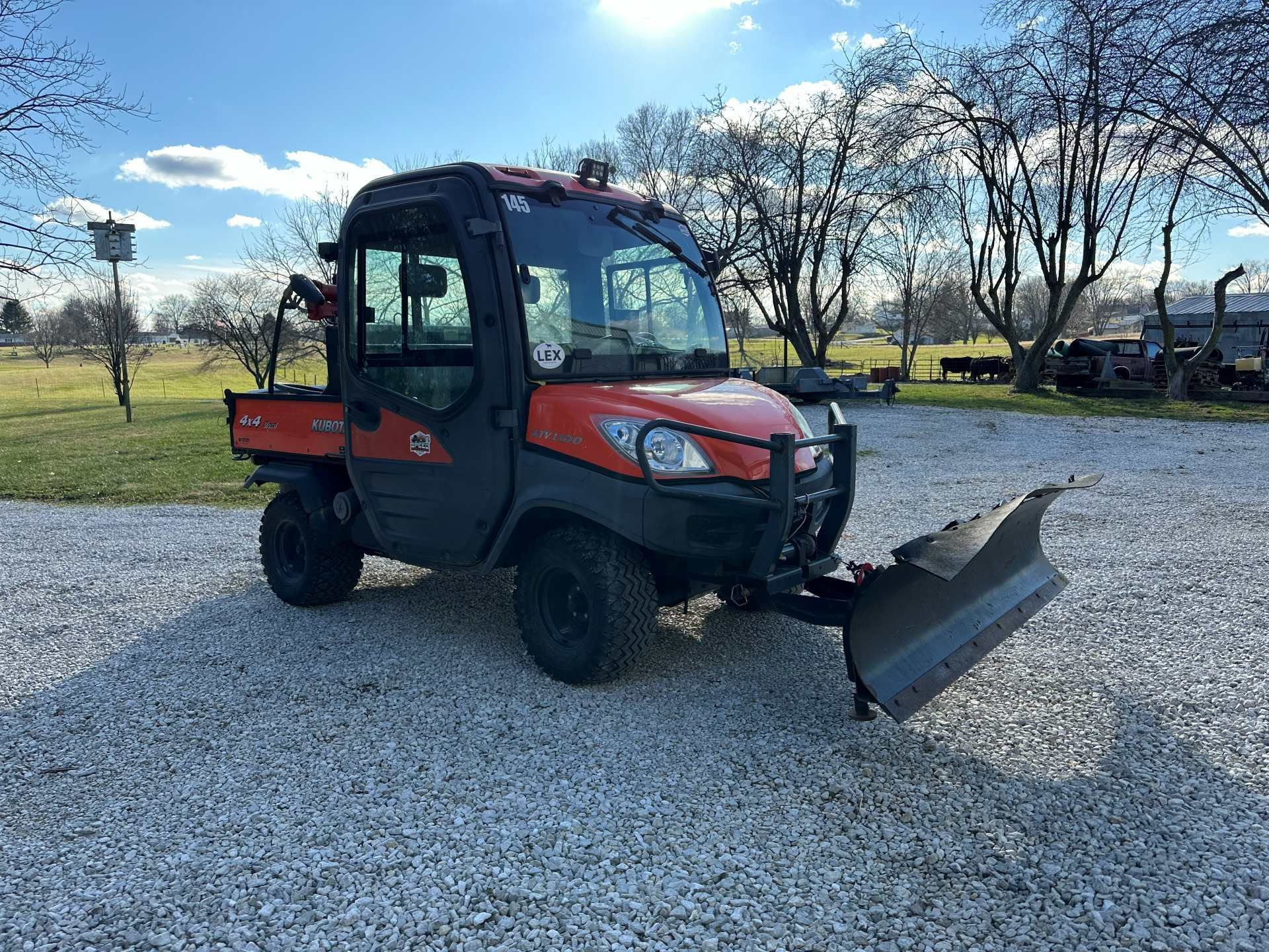 An orange Kubota RTV utility vehicle with a snow plow blade attached, parked on a gravel lot under a sunny sky.