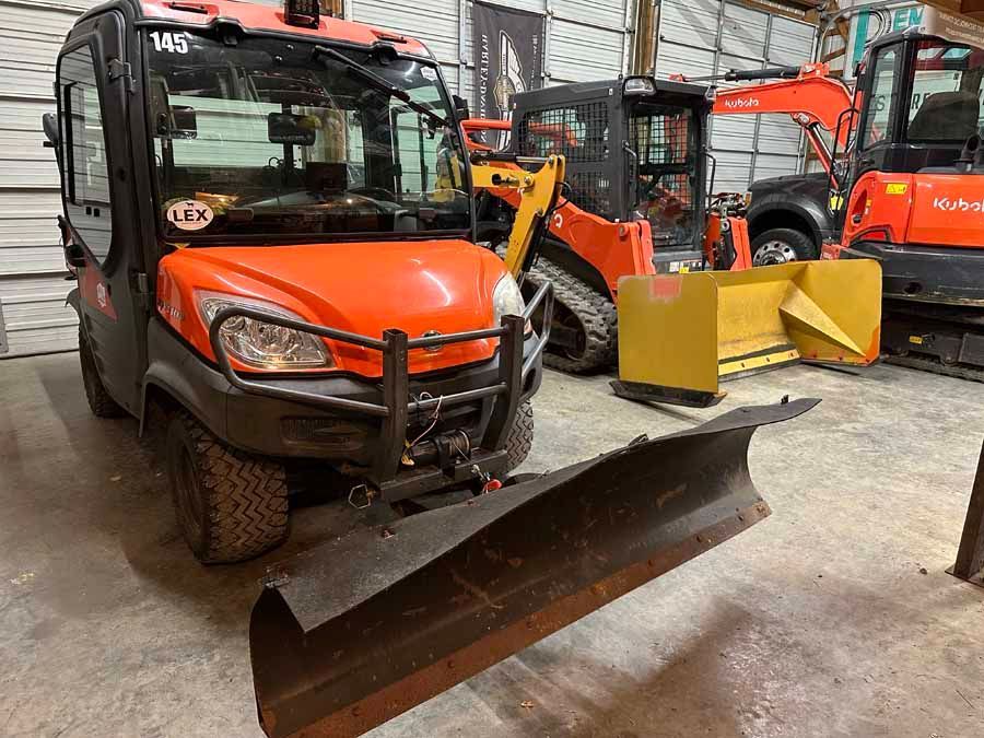 An orange Kubota utility vehicle with a mounted snow plow parked indoors next to a skid steer loader and mini excavator.