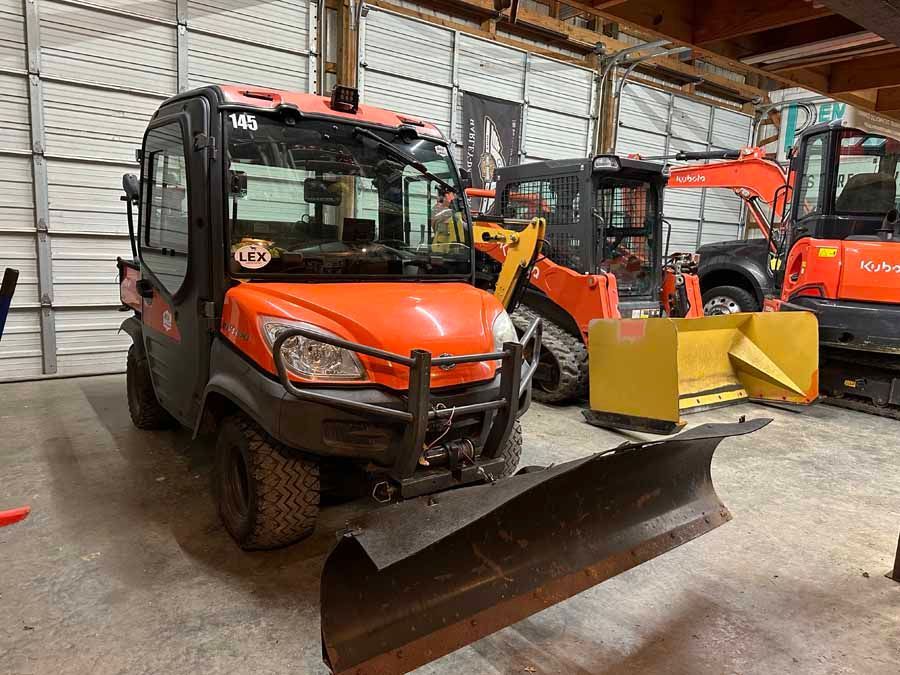 An orange Kubota RTV with a snow plow blade parked in a warehouse next to a skid steer and an orange excavator.