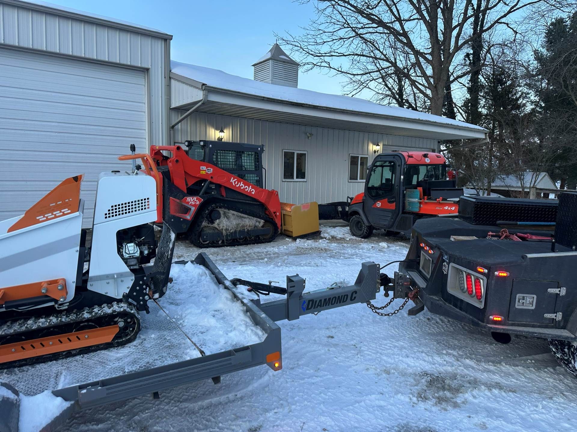 A white and orange skid steer on a trailer, with an orange loader parked behind it in front of a white building in snow.