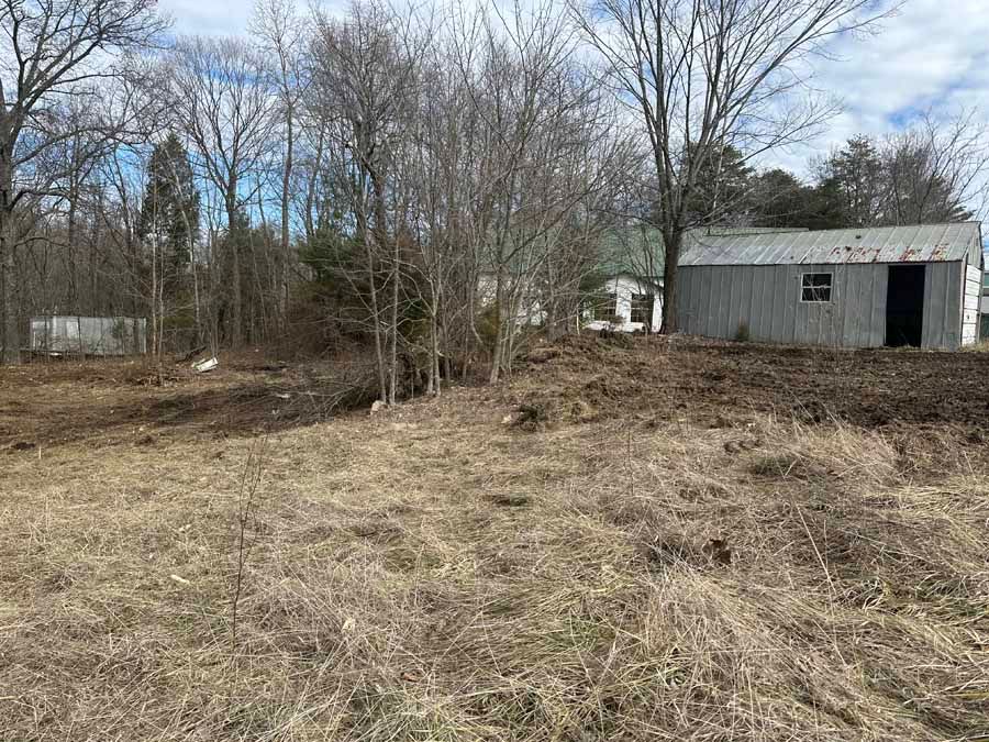 A view of a muddy, cleared lot with dry grass in the foreground and a metal shed on the right near sparse, leafless trees.