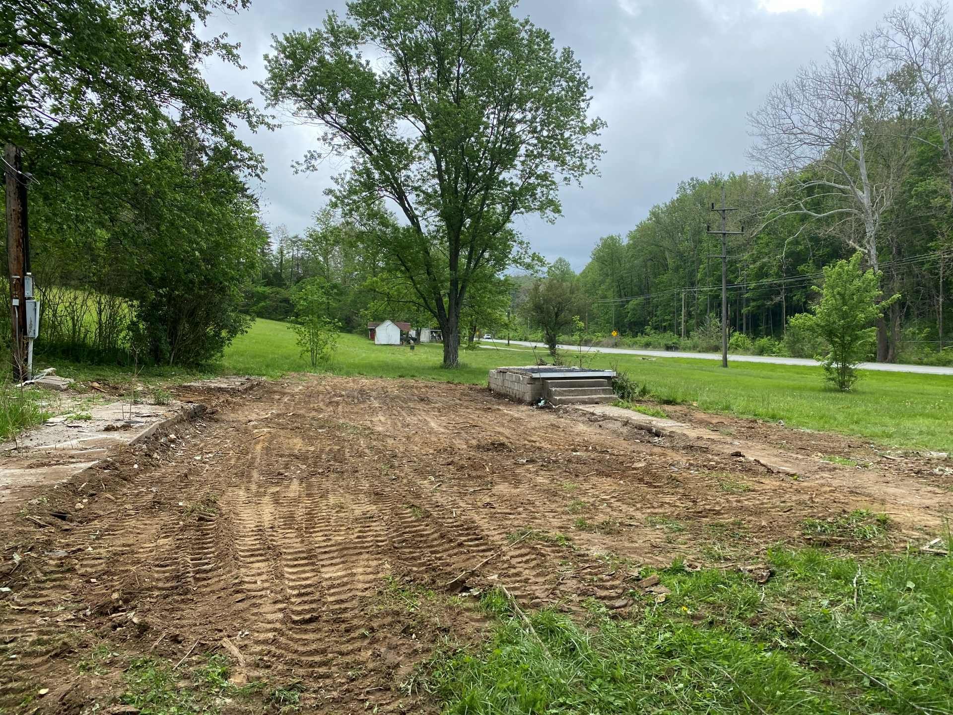 A cleared, dirt-covered lot with tire tracks, set in a grassy landscape with trees under an overcast sky.