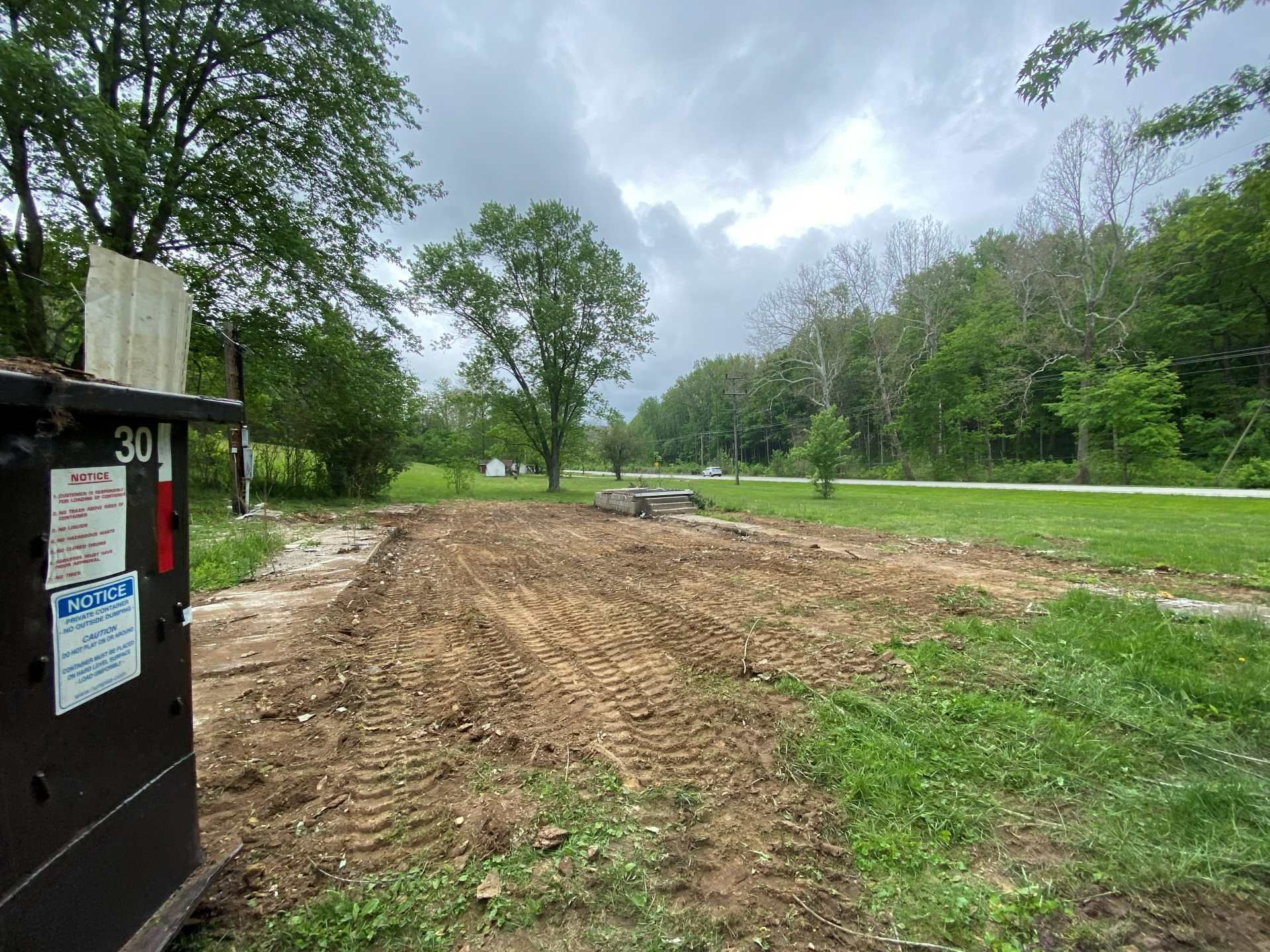 A dark dumpster sits in the foreground beside a cleared, dirt-covered construction site under a cloudy sky.