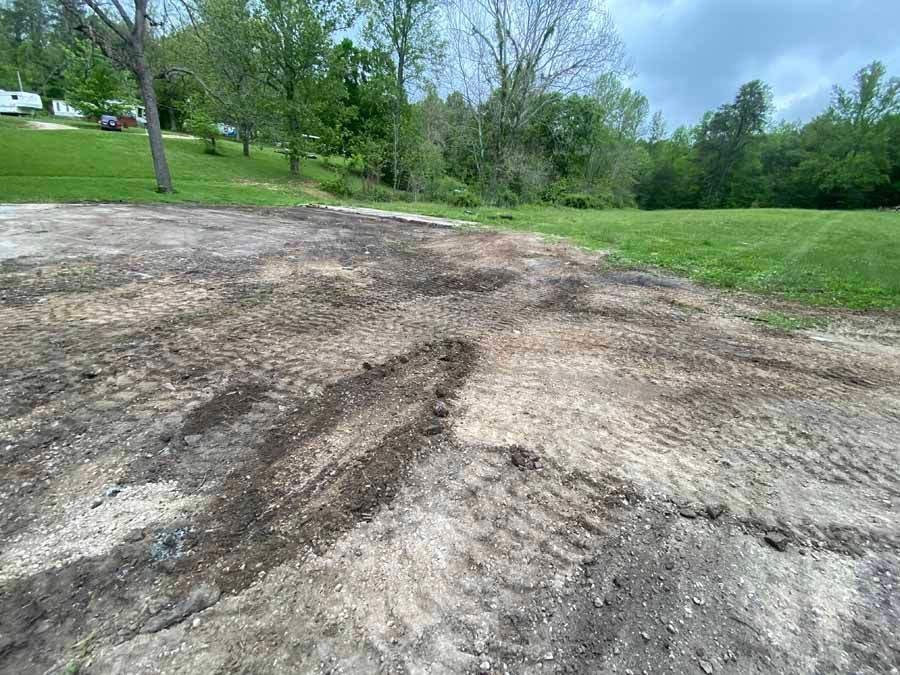 A patch of cleared, uneven dirt ground with tire tracks in a grassy, rural setting with trees under a cloudy sky.